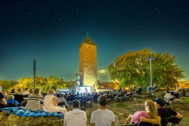Thomas Ziegler, Stadt Halle (Saale) Abendveranstaltung im Freien mit vielen Zuschauern, die auf Liegestühlen und Decken sitzen und einem beleuchteten Turm im Hintergrund, der von Bäumen umgeben ist.