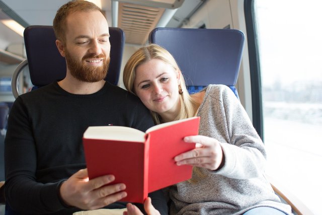 Martin Neuhof Zwei Personen auf einem Zug sitzen gemeinsam eng beieinander und blättern gemeinsam in einem roten Buch. Die Frau zeigt auf eine Seite, der Mann blickt aufmerksam in das Buch.