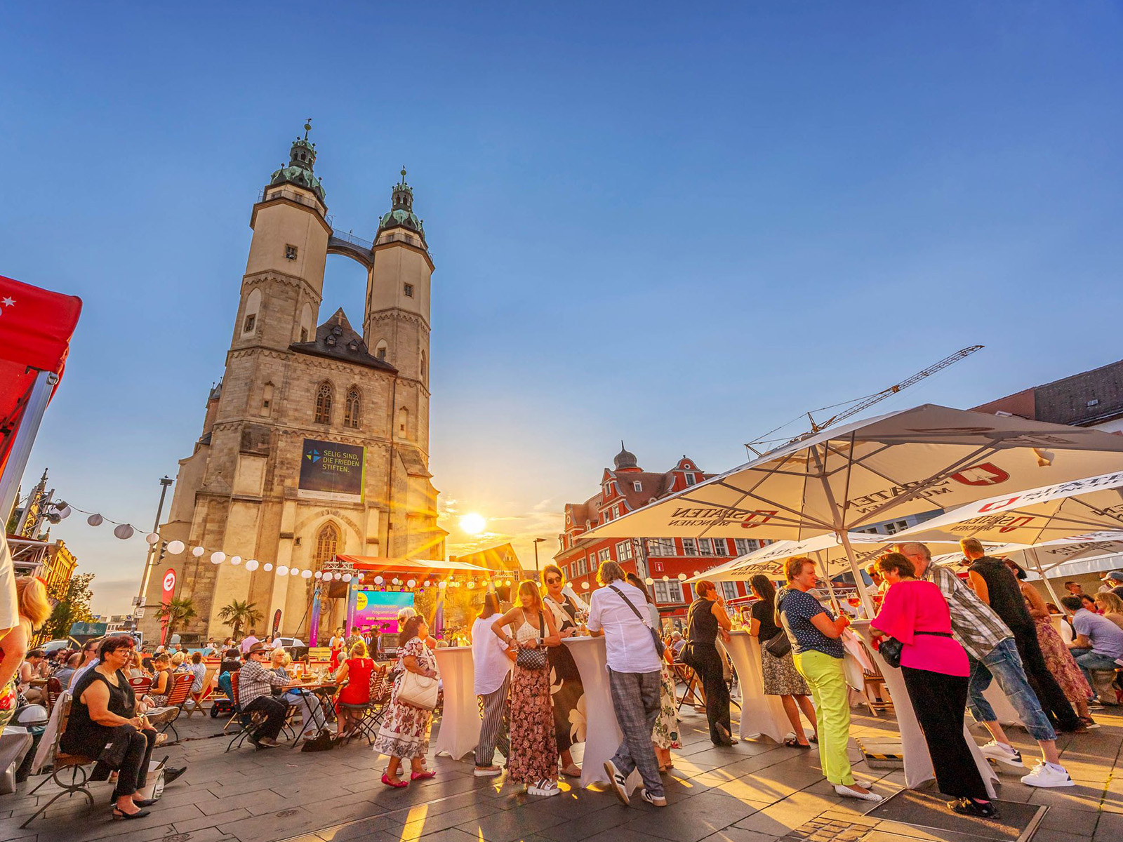 Thomas Ziegler, Stadt Halle (Saale) Abendliche Markt- oder Festplatzszene mit beleuchteter historischer Kirche im Hintergrund, zwei markanten Türmen und Kuppel. Vorne viele Menschen, die an Ständen mit bunten Markisen essen, trinken und sich unterhalten. Lebhafte Atmosphäre bei Sonnenuntergang.